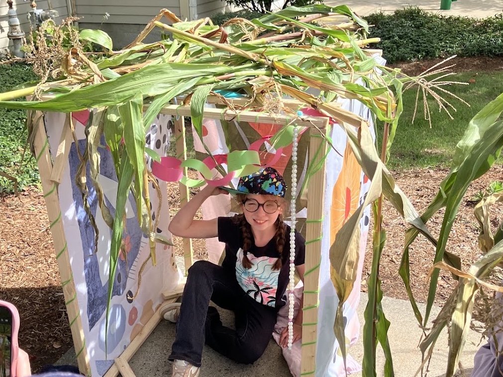student in small sukkah with cornstalks on top and colorful sheets as walls