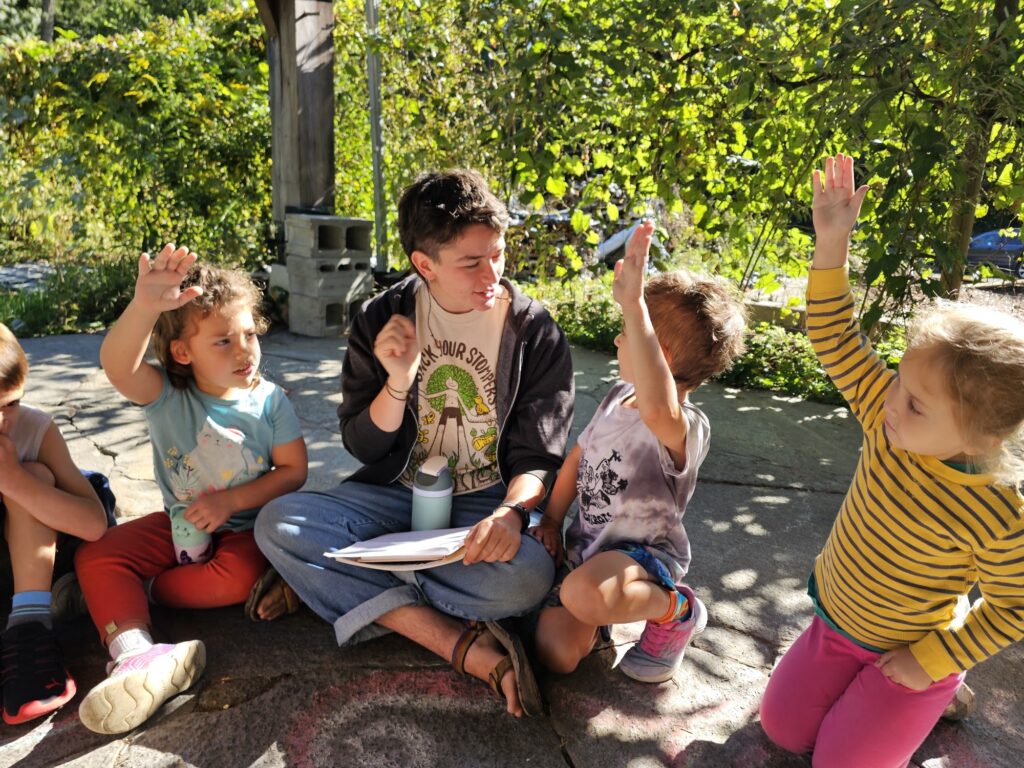 three kids and one teacher sitting together, kids are raising hands. They are outside sitting on the ground. Teacher is smiling